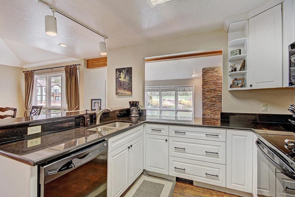 Modern kitchen with white cabinets, black countertops, stainless steel appliances, and a double sink. The space includes an adjacent dining area with large windows and beige curtains.