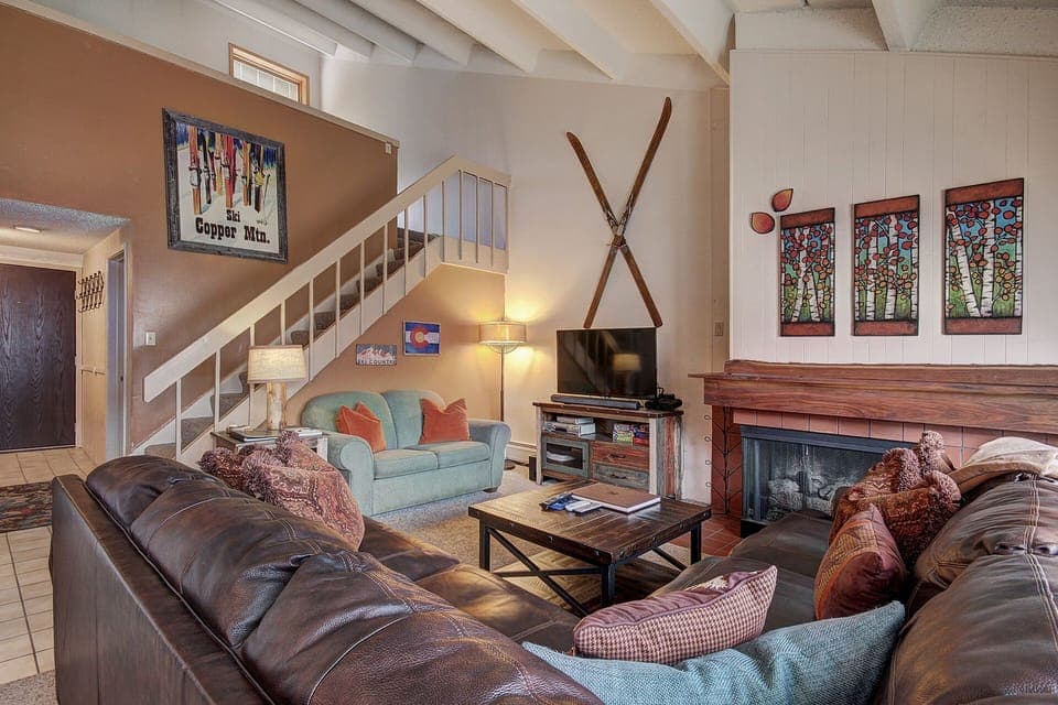 Living room with a staircase, a sofa, a loveseat, a coffee table, a flat-screen TV, and a fireplace. Wall decor includes crossed skis and art pieces. The room has a cozy, rustic feel.