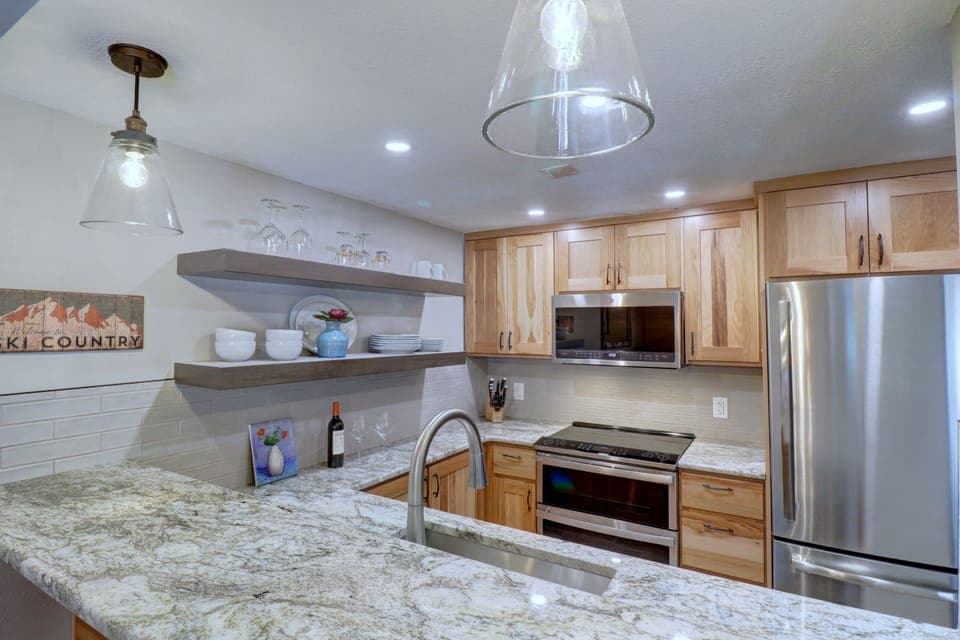 Modern kitchen with granite countertops, stainless steel appliances, wooden cabinets, open shelving with dishes, and pendant lighting. A "Ski Country" sign hangs on the wall.