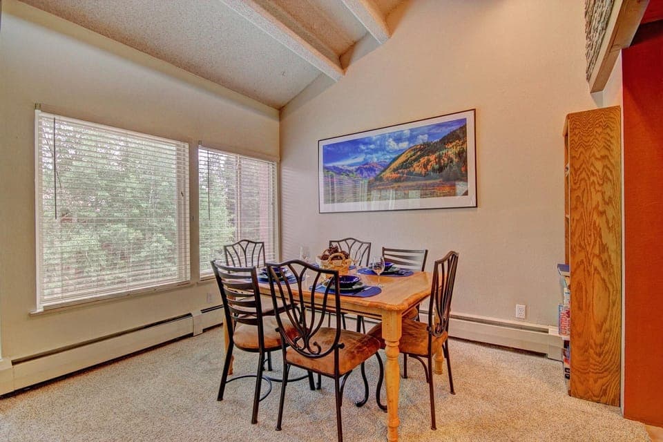 A dining room with a wooden table set for four with blue place settings. The room features large windows, a landscape painting on the wall, and wood and metal chairs.