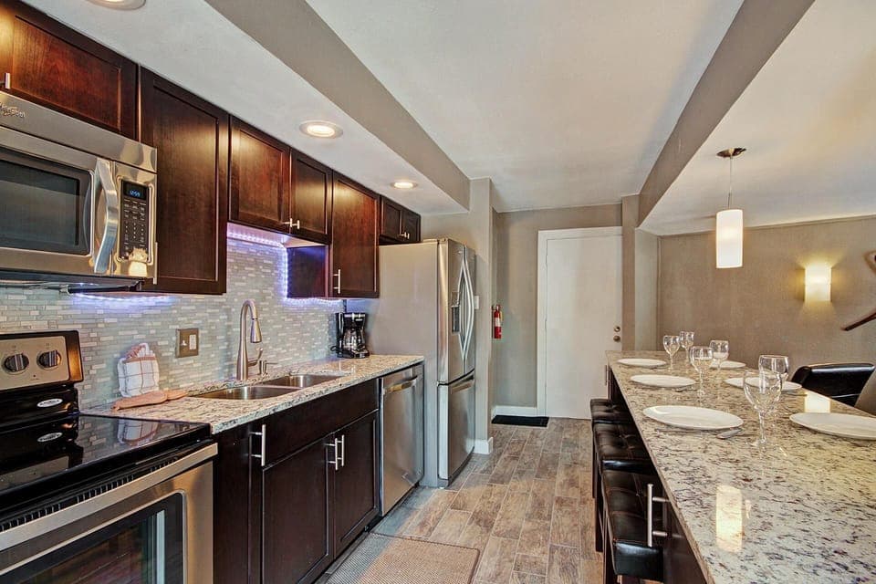 A modern kitchen with stainless steel appliances, dark wood cabinets, granite countertops, and a kitchen island with seating and place settings. A pendant light hangs above the dining area.