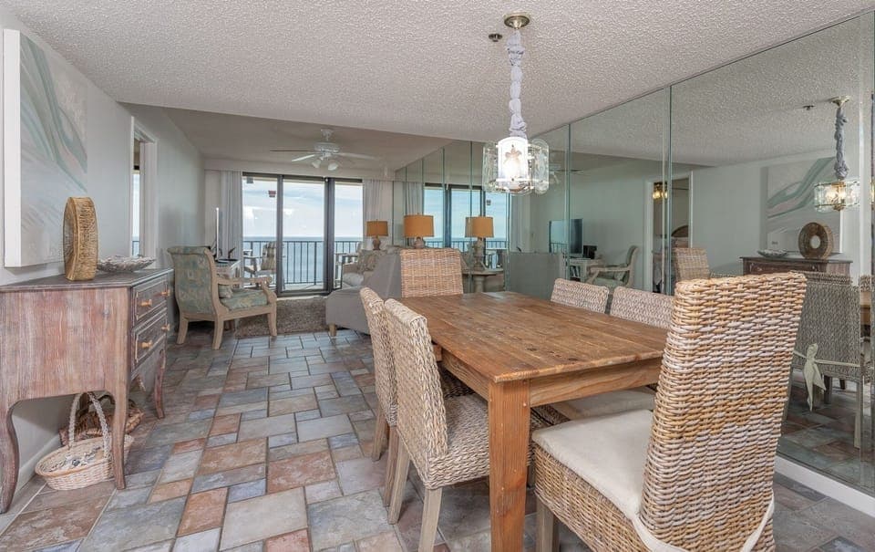 The dining area features a rustic wooden table with woven chairs, with a beautiful beach view visible through large windows