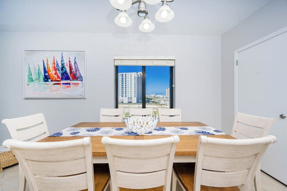 Gather around the inviting dining table, accented by a colorful sailboat painting and a view through the window