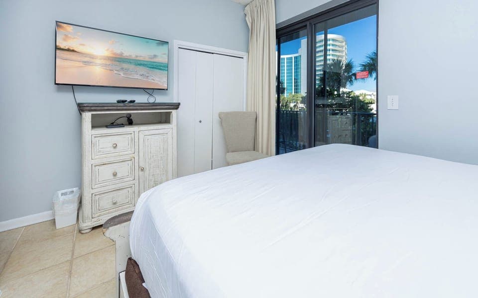 Relax in this comfortable guest bedroom featuring a large TV, a cozy chair, and natural light from the balcony doors