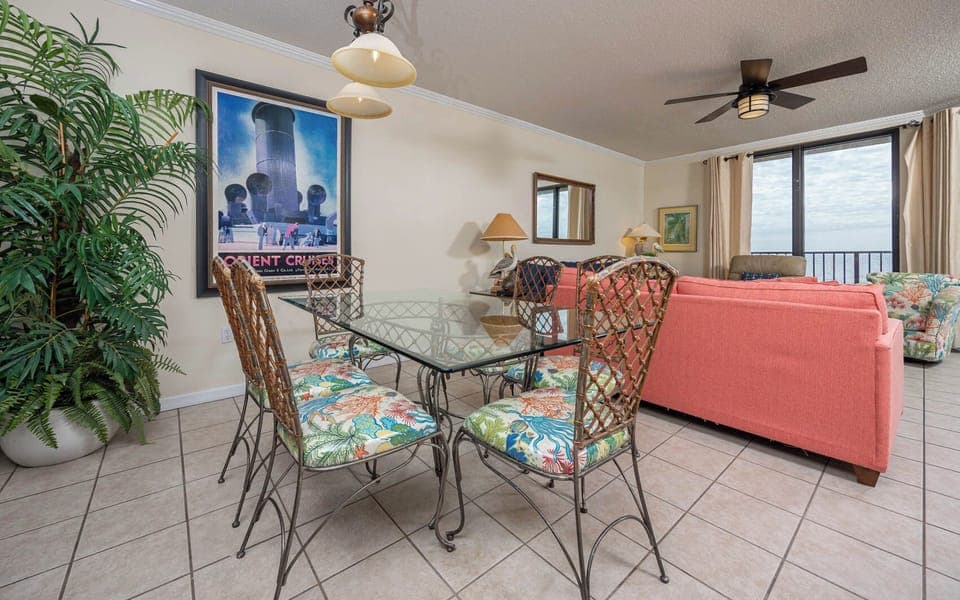 Gather around the glass dining table adorned with vibrant chairs, perfect for meals with a view of the coastline