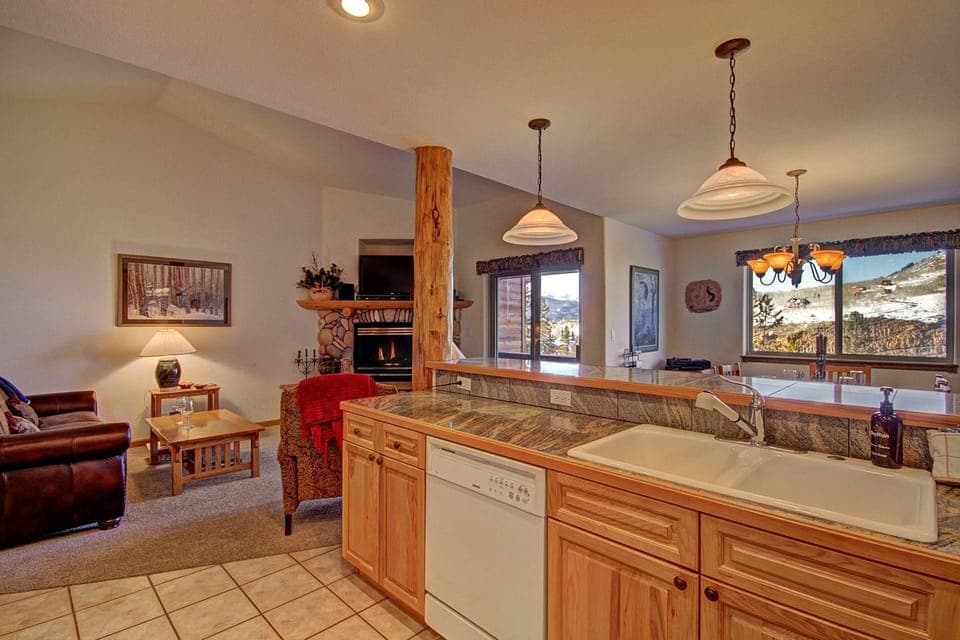 A cozy open-plan living room and kitchen area with wooden cabinetry, modern appliances, a leather sofa, a stone fireplace, large windows, and mountain views.
