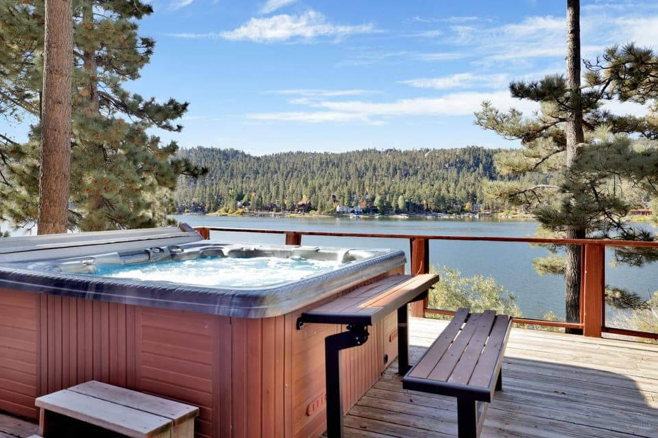 A bubbling hot tub sits on a wooden deck, offering a stunning view of a serene lake and pine-covered hills. This peaceful retreat captures the essence of a perfect mountain getaway under a blue sky.