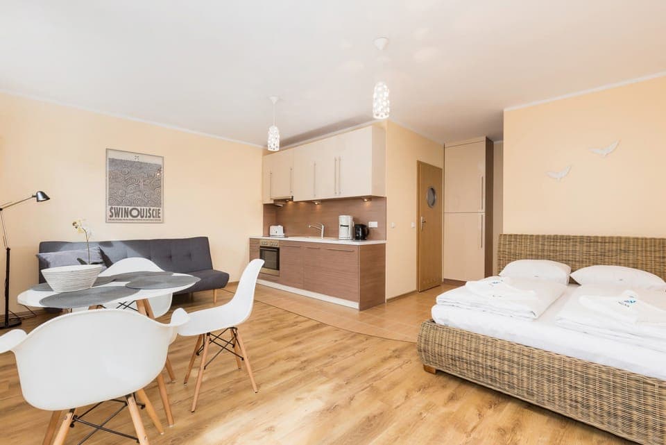 The room features a queen-size bed in an open-plan space. There’s a round dining table with two modern chairs, a compact, sleek design, and a TV mounted on the wall.
