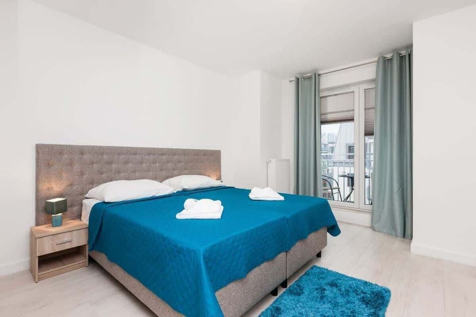 A cozy bedroom with a blue bedspread on a queen-sized bed, flanked by a simple headboard. In the background, there is a wardrobe with mirrored doors and a glimpse of the adjoining area.