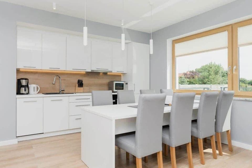A modern kitchen and dining area with a white, minimalist design, featuring a spacious dining table for six and a sleek kitchen setup with wooden accents.
