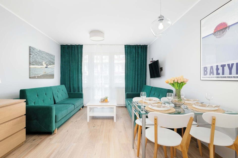 A modern living room featuring a green sofa, a dining table with chairs, and large windows with green curtains.