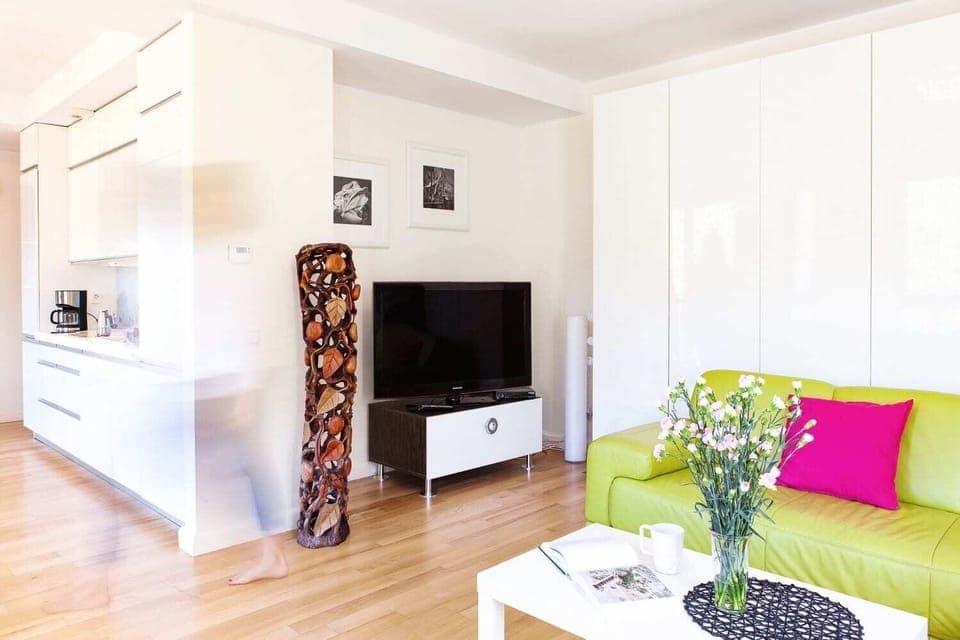 Another angle of the living room showing a white console and green sofa with pink and purple pillows. A modern, minimalist aesthetic is enhanced by colorful decor and soft lighting.
