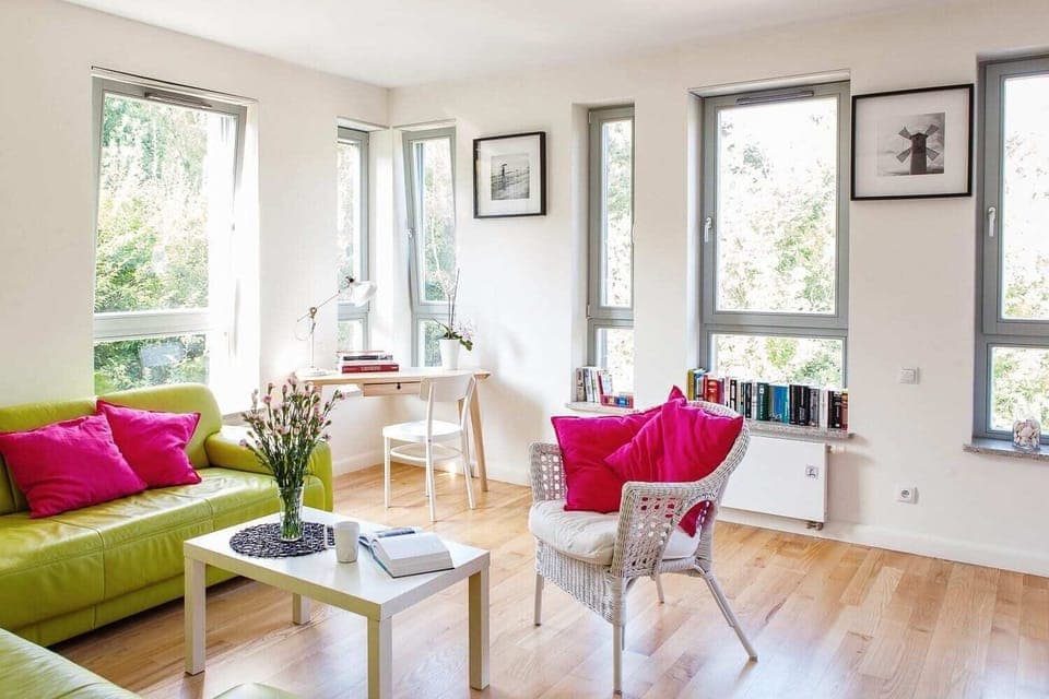  A spacious, light-filled living room with a green sofa, pink cushions, and a glass coffee table. Large windows allow natural light to brighten the space, and a small dining table is set up near the sofa.