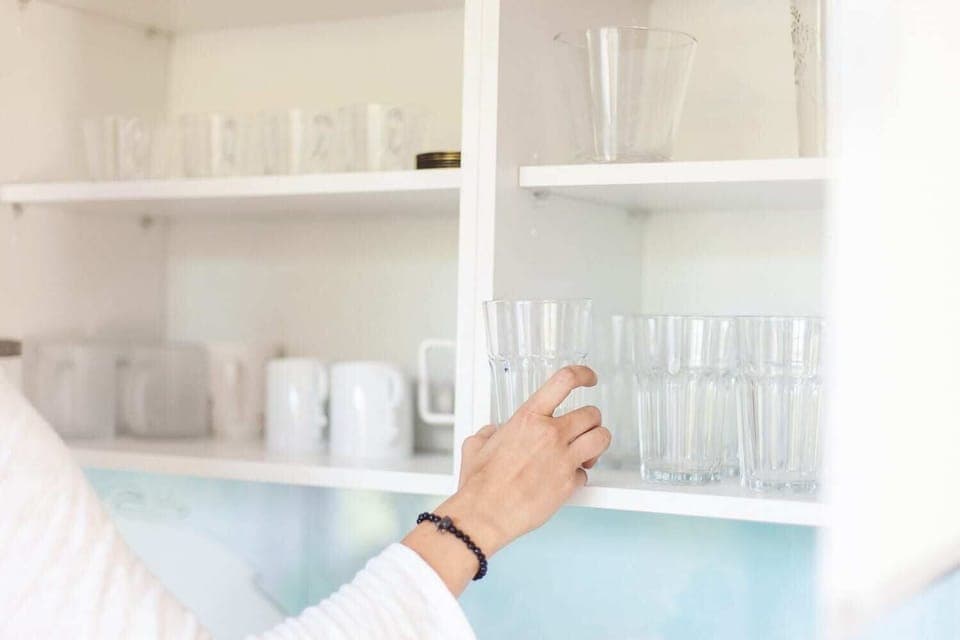  A close-up of a person reaching for glassware inside a white kitchen cabinet. The cabinet is organized, with clear glass cups and white plates neatly arranged.