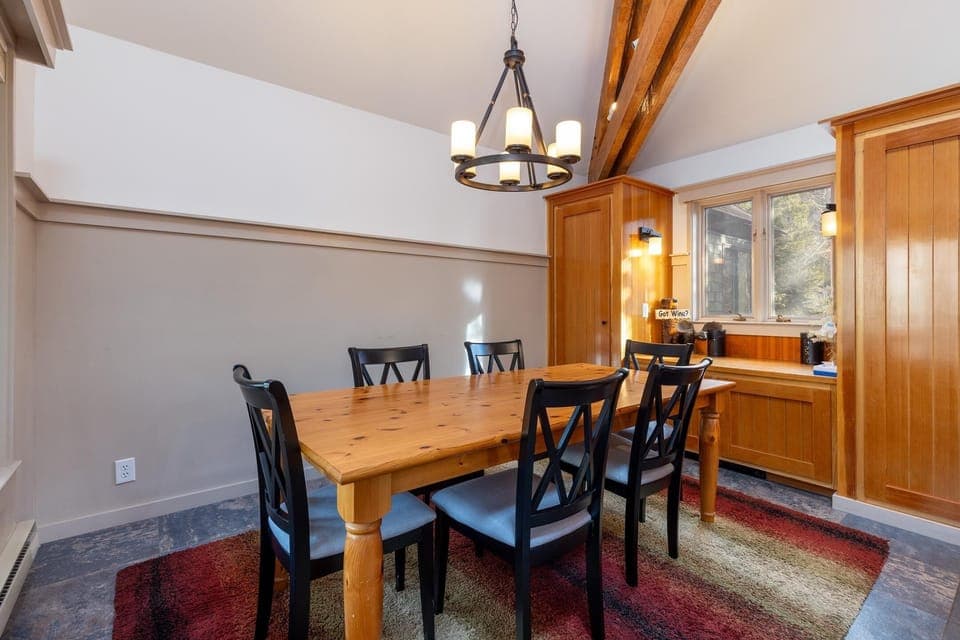 A dining room with a wooden table and six black chairs, a chandelier overhead, and natural light coming through a window. The room has a two-tone wall, exposed wooden beams, and an area rug.