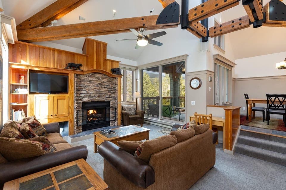 A spacious living room with wooden beams, a stone fireplace, sofas, a television, and large windows overlooking a forest. A dining area with a table and chairs is visible in the background.