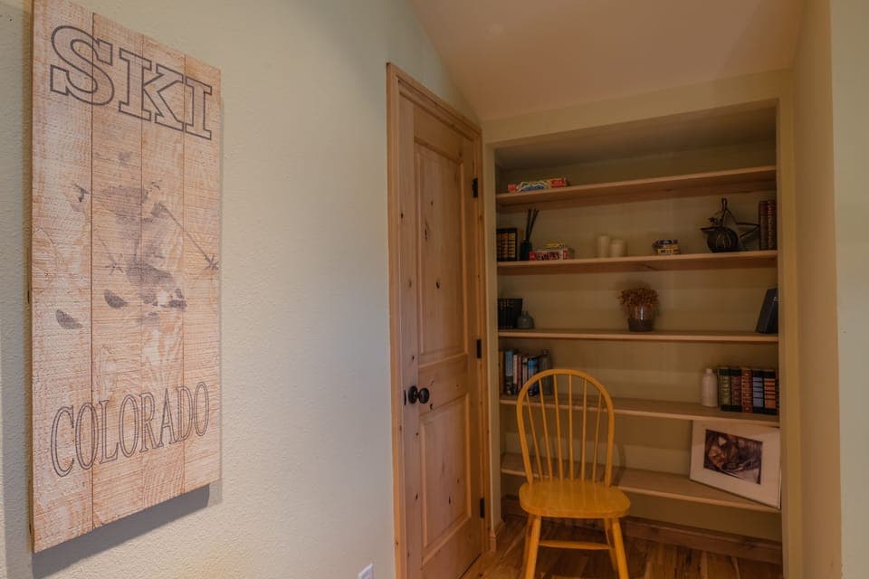 A small room with a wooden door and built-in shelves. On the shelves are books, small decorative items, and a framed photo. A wooden chair is placed in front of the shelves. A "Ski Colorado" sign hangs on the wall.