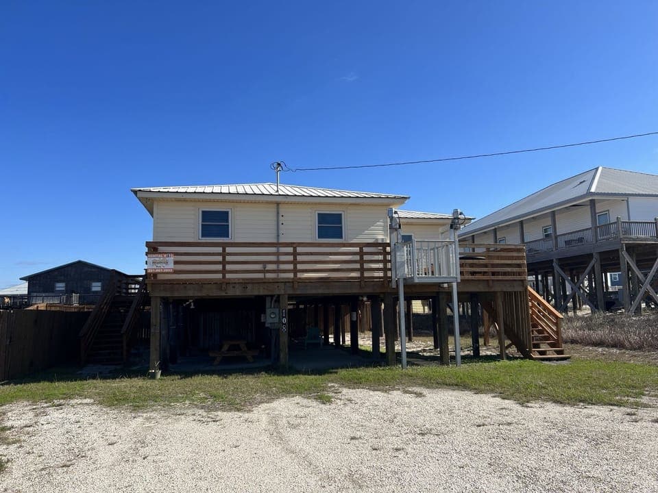 Waterfront property elevated on stilts with wraparound deck and stairs, situated in coastal neighborhood.