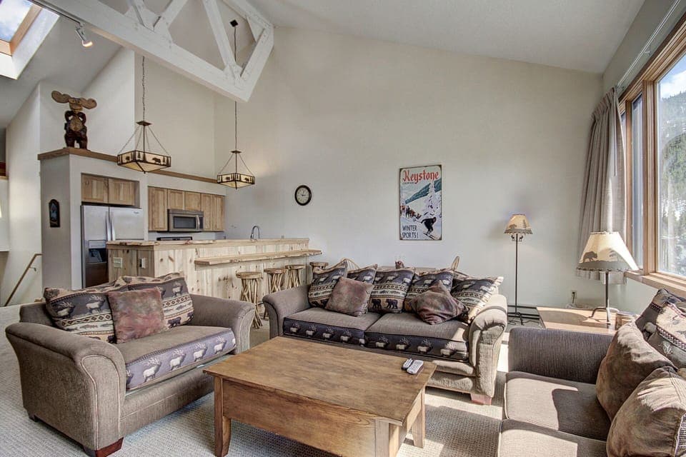 A cozy living room features two sofas, a coffee table, a kitchen area with wooden countertops, and a bar counter. The room is decorated with a bear sculpture and posters, with large windows allowing natural light.