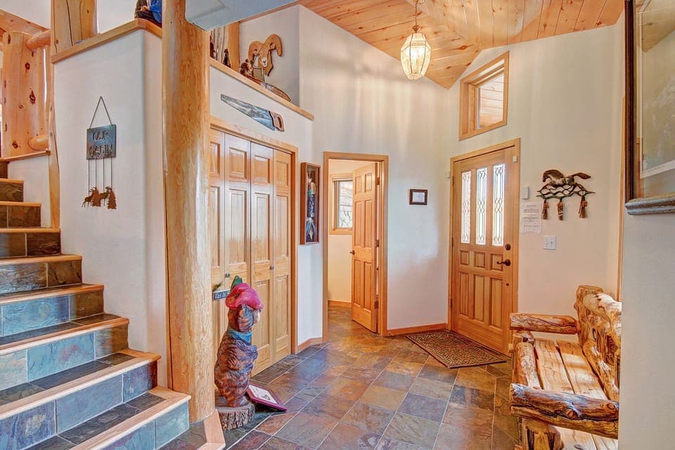 Entryway featuring wooden doors, a rustic bench, a staircase with decorative posts, and various carved wood decorations. There is a chandelier and natural light illuminating the space.