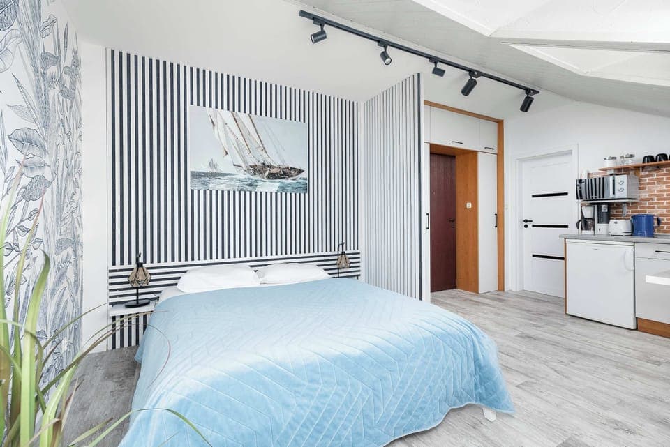 Close-up of the comfortable double bed on the left, the kitchen area with white furniture is visible. 