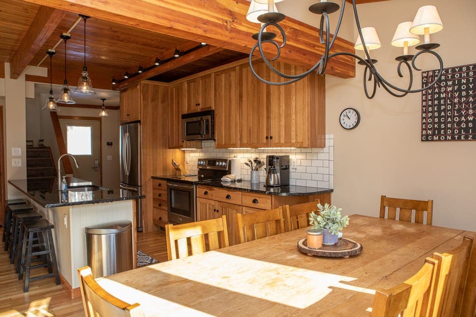A kitchen with wooden cabinets, stainless steel appliances, a center island with bar stools, and a dining table. A chandelier hangs above the table, and a clock and word art are on the wall.
