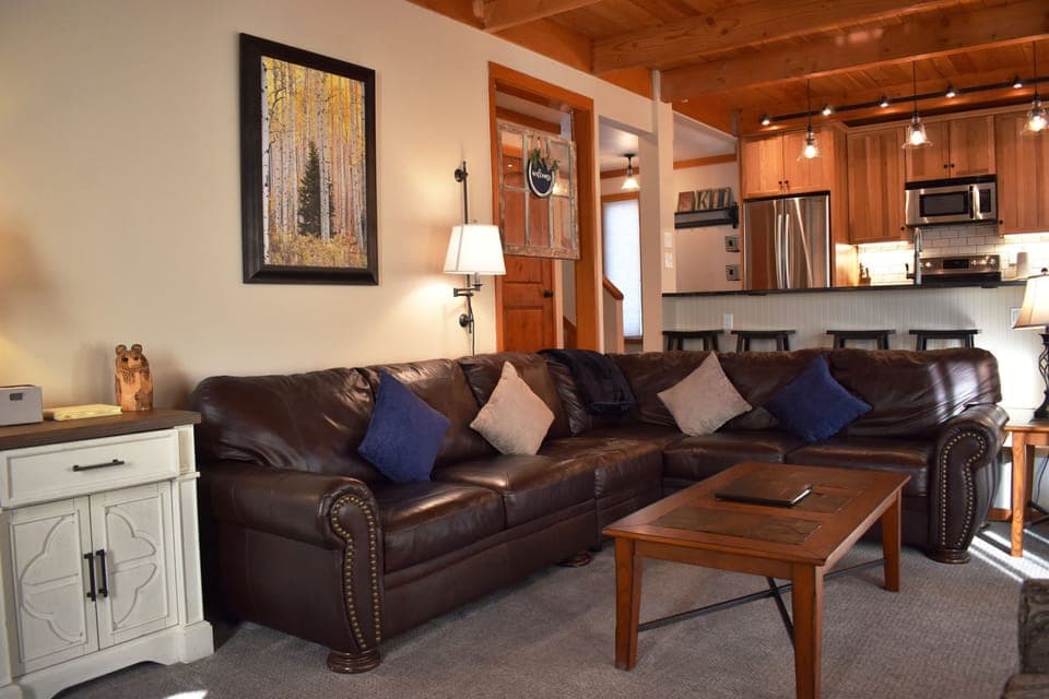 A cozy living room with a brown leather sectional sofa, decorated with pillows. There is a framed painting on the wall, a wooden coffee table, and a view of the kitchen in the background.