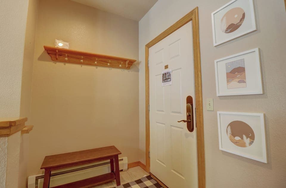Entryway features a white door, wooden bench, coat rack with hooks, and framed artwork on the wall. The floor has a checkered rug and beige walls.