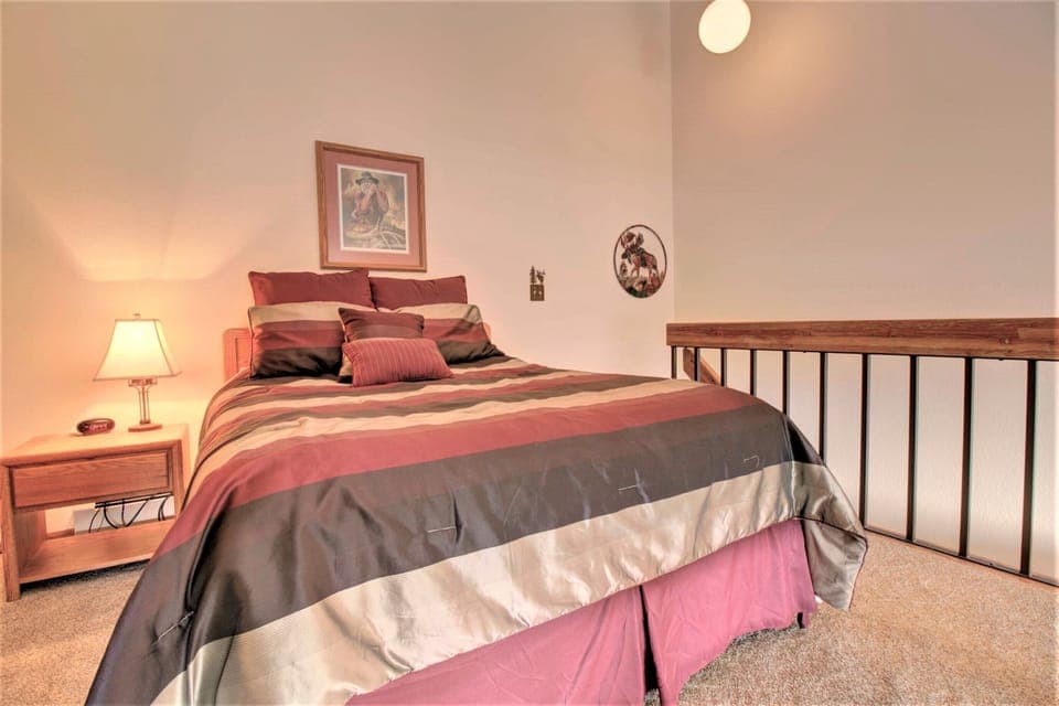 A bedroom with a neatly made bed featuring a striped comforter, a nightstand with a lit lamp, and framed art on the wall. The room has a high ceiling and railing beside the bed.