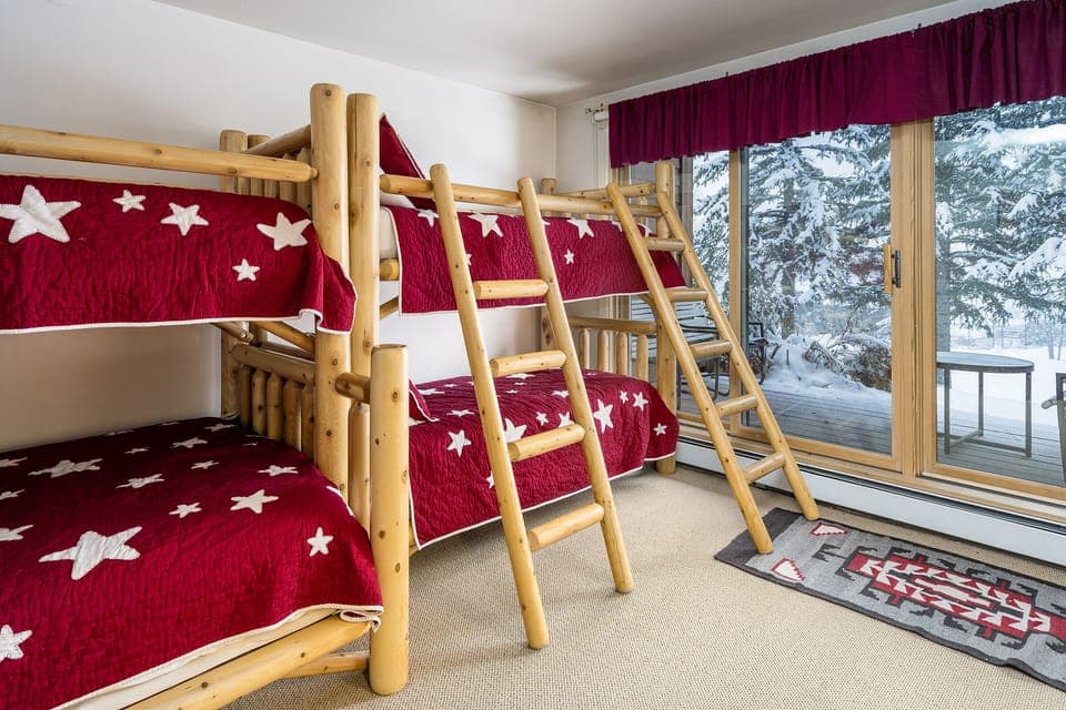 A room with wooden bunk beds featuring red star-patterned bedding. The space has a sliding glass door leading to an outdoor area with snow-covered trees. A rug with a geometric design lies on the floor.