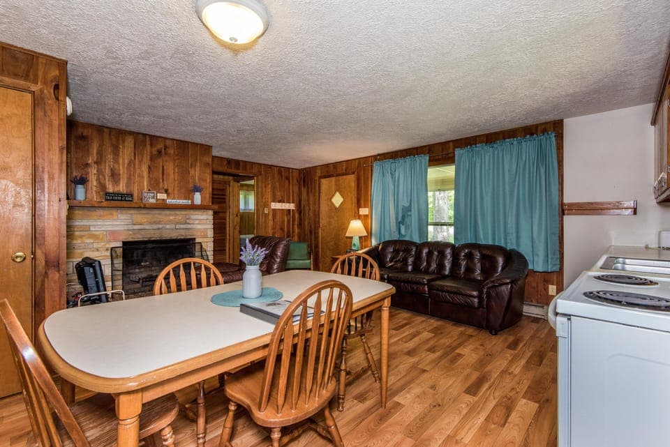 Dining table and wooden chairs sit beside a stone fireplace and a dark leather sofa with blue curtains.