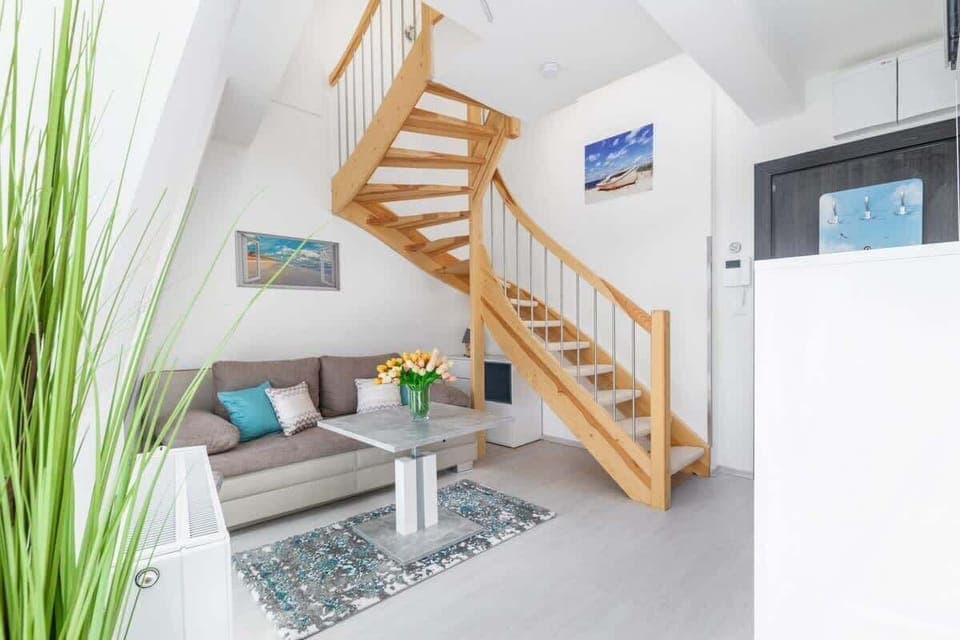 A cozy living room with a staircase leading to an upper floor. The room is decorated with plants and has a bright atmosphere.
