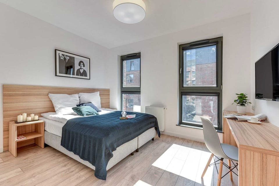 A bright bedroom featuring a comfortable bed with blue bedding, a small desk, and large windows providing natural light. The space is modern and functional, ideal for work or relaxation.