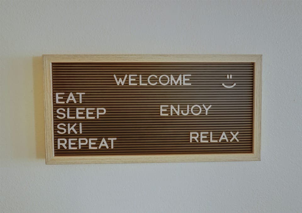 A wooden-framed sign with white letters reads, "WELCOME. EAT SLEEP SKI REPEAT ENJOY RELAX" on a brown background. There is a smiley face next to the word "WELCOME.