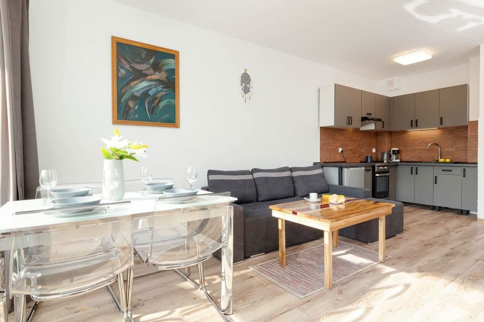 A modern living room with a gray sofa, a wooden coffee table, and a TV unit, connected to an open-plan kitchen.