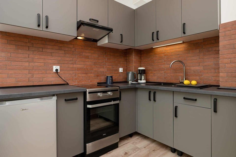A compact, modern kitchen featuring gray cabinets, a red brick backsplash, and black countertops.