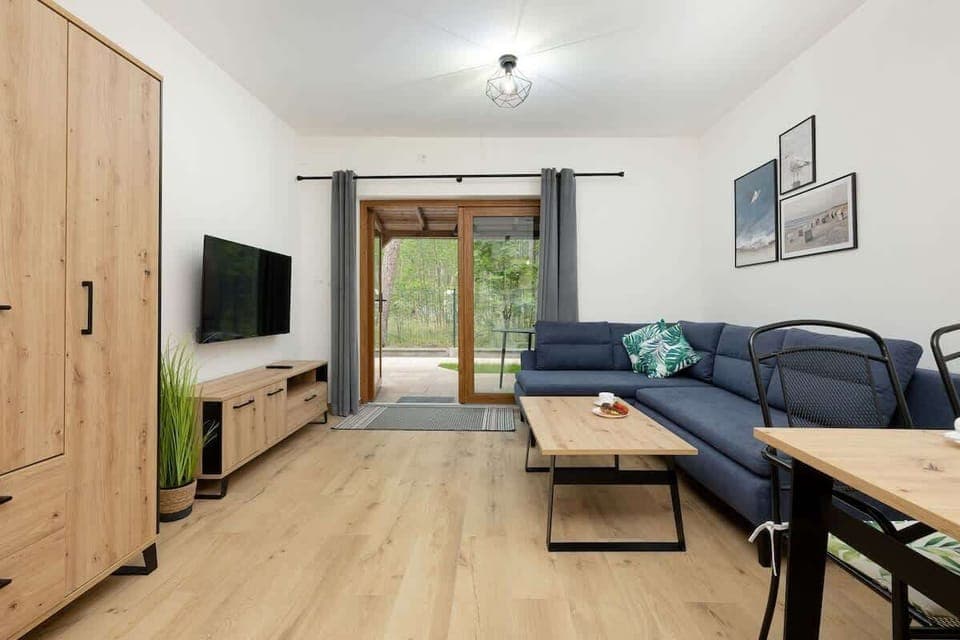 A cozy space with wooden flooring, a large TV mounted on the wall, a light wooden TV stand, and a comfortable navy-blue sofa with a small wooden coffee table. The room is bright, featuring large windows that let in natural light.