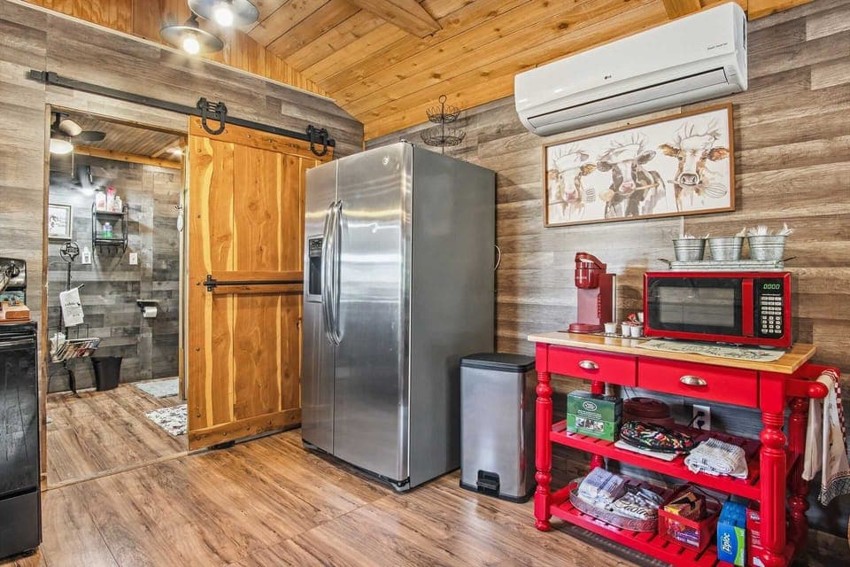 A interior view of the cabin's kitchenette area, featuring a large stainless steel refrigerator, a bright red utility cart with a microwave and coffee station, and a sliding wooden barn door that opens into a modern bathroom.