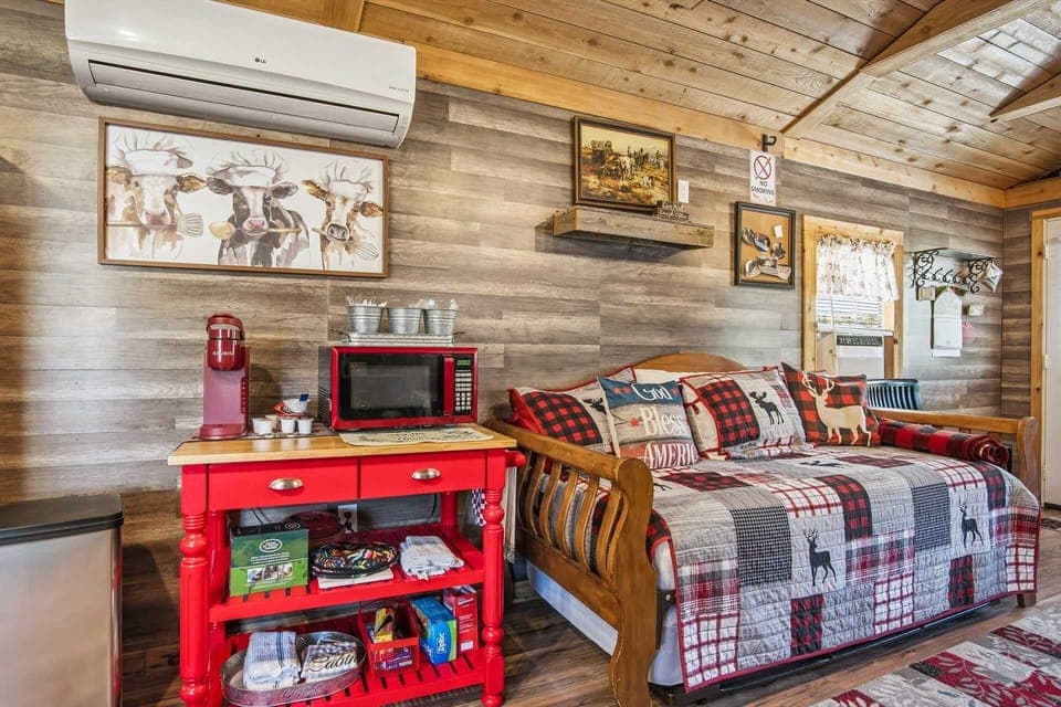 A cozy corner of the cabin featuring a bright red kitchen cart equipped with a microwave and coffee maker, situated next to a rustic wooden daybed with lodge-inspired patchwork bedding.