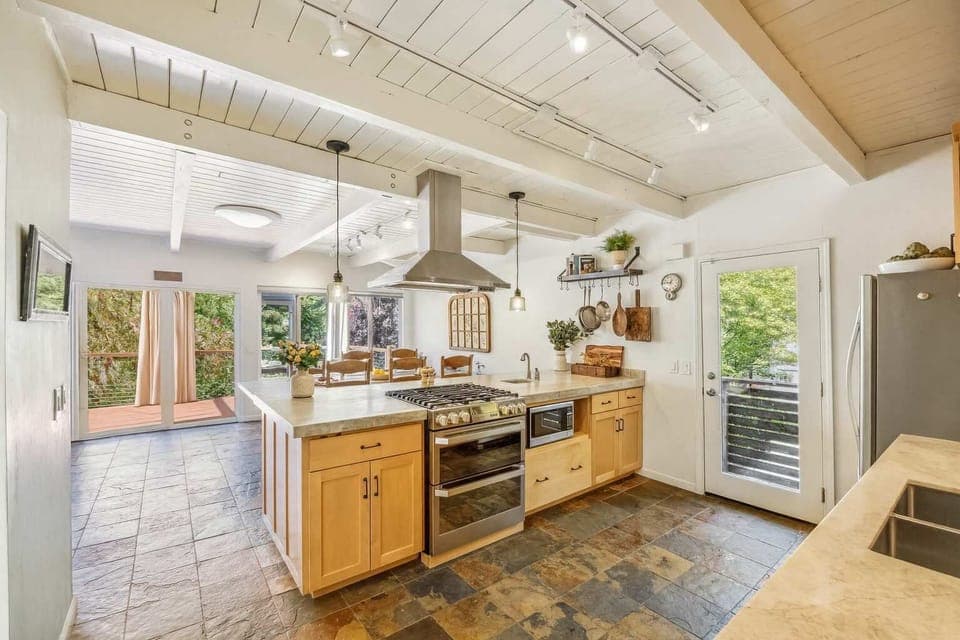 A warm, beautifully lit kitchen with natural wood cabinets and a large central island. Sliding doors open to the deck, making cooking, dining, and entertaining feel effortless.