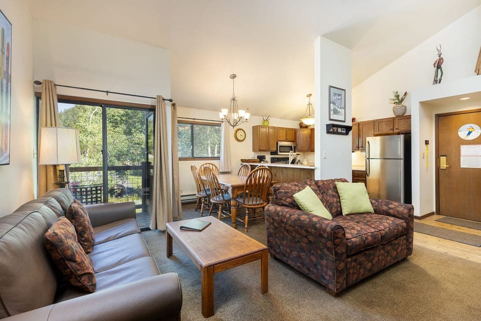 A cozy living room and kitchen area featuring a sofa, an armchair, a wooden coffee table, a dining table with chairs, a modern kitchen, and large windows with a view of greenery.