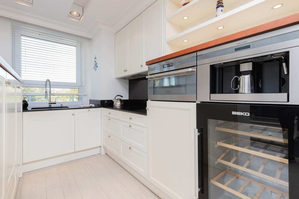 A sleek kitchen featuring white cabinets, a built-in oven, and a wine cooler, all complemented by a black countertop.

