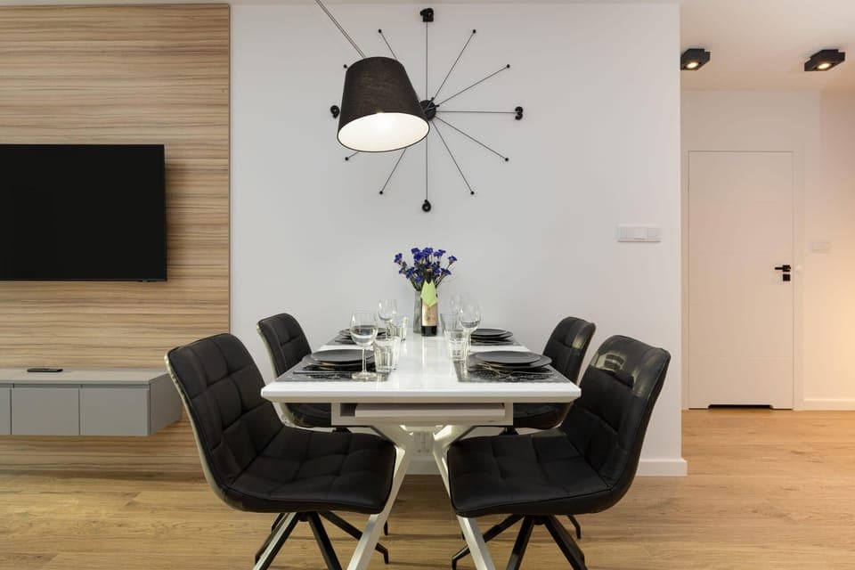 Dining area with white table and black chairs over which hangs a modern large clock.