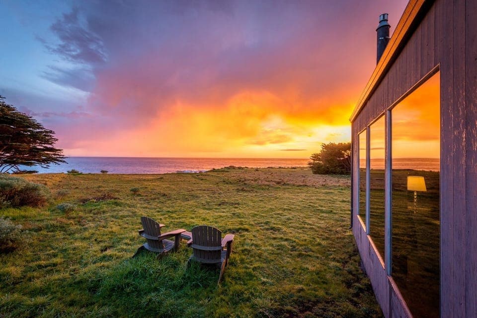 Two Adirondack chairs face the Pacific, where evening light and color reflect across the home’s modern glazing, marking the quiet transition from day to dusk.
