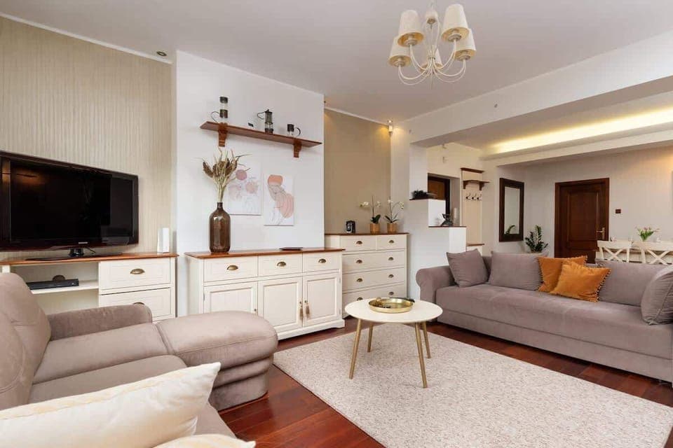  Bright living room featuring a large TV unit, white furniture, and decorative elements such as a chandelier and plants. Wooden flooring adds warmth.
