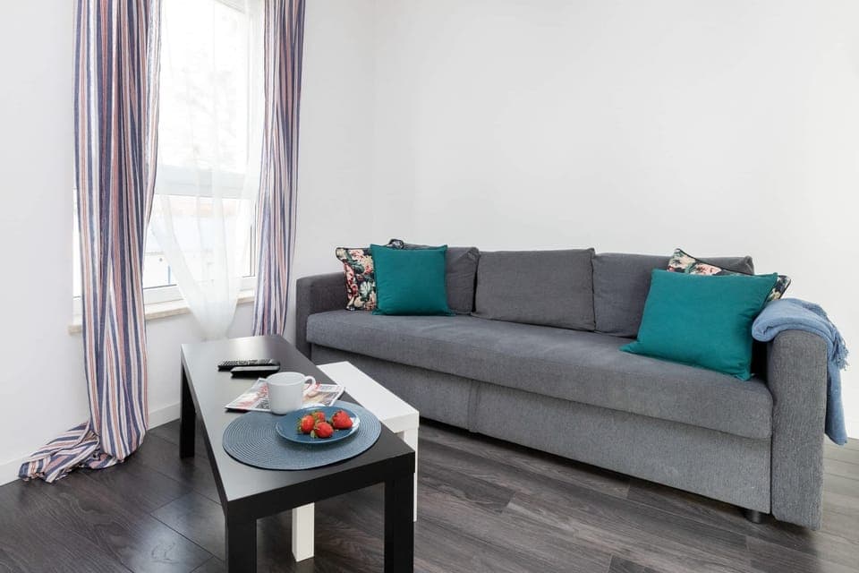 A cozy living room view with gray sofas, teal cushions, a small table, and bright natural light from the windows.