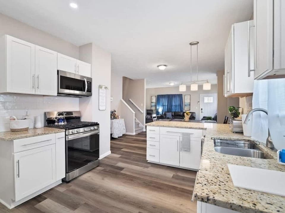 White kitchen boasts granite counters, island workspace under pendant lights, stainless gas range, and a sleek sink. Open layout flows seamlessly into the living area—perfect for cooking & socializing! ✨