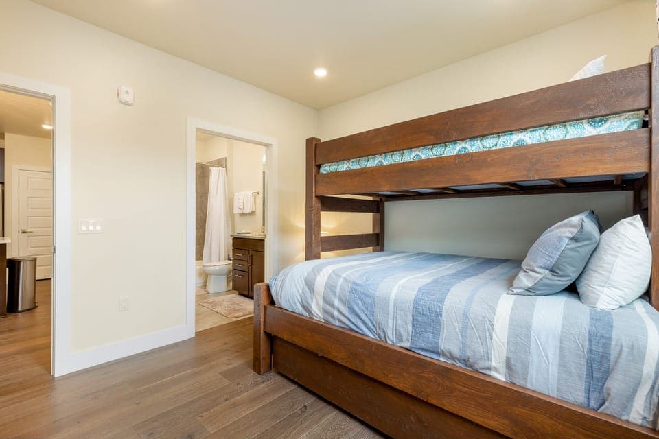 A bedroom with wooden bunk beds, each made up with blue and white linens, next to an open door leading to an adjacent bathroom. The walls and ceiling are off-white, and the floor is wooden.