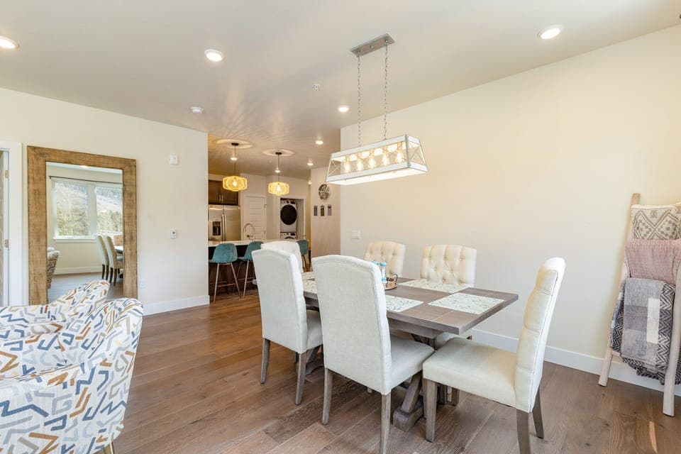 A modern dining room with a rectangular table, six padded chairs, a pendant light, and an adjacent open kitchen. Light wood flooring and white walls create a bright, airy atmosphere.