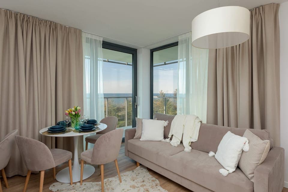 This modern living room features a comfortable sofa facing a large window, offering direct access to a balcony. The mix of soft textures and natural light creates a welcoming environment with a great connection to the outdoor space.
